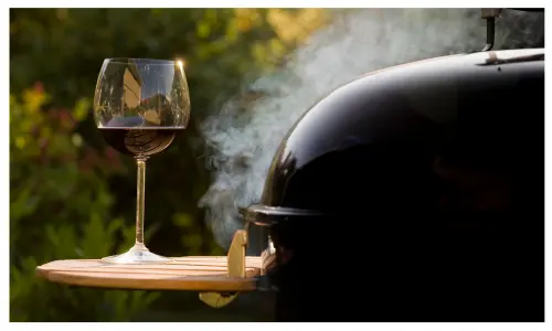 A steaming black domed barbecue with a wooden shelf on which there is a glass of red wine