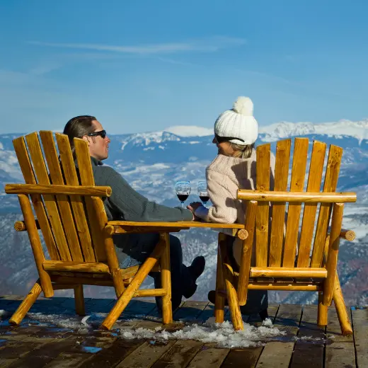 Man and a woman wearing sunglasses and warm clothing sitting on wooden chairs on top of a mountain each holding a glass of red wine