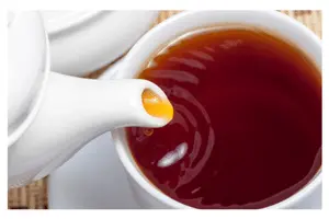 Spout of a white teapot from which black tea is being poured into a white cup