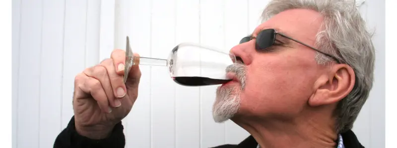 A man with grey hair and a beard wearing dark glasses and drinking some red wine