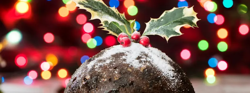 A typical British Christmas pudding with a sprig of holly on top and colourful fairy lights in the background