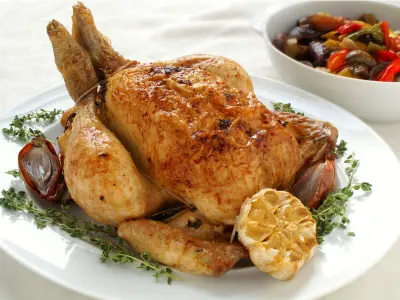 A cooked chicken on a white platter with some roated garlic and on the side a white dish filled with ratatouille