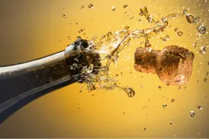Top of a bottle of sparkling wine with the cork exploding out against a yellow background