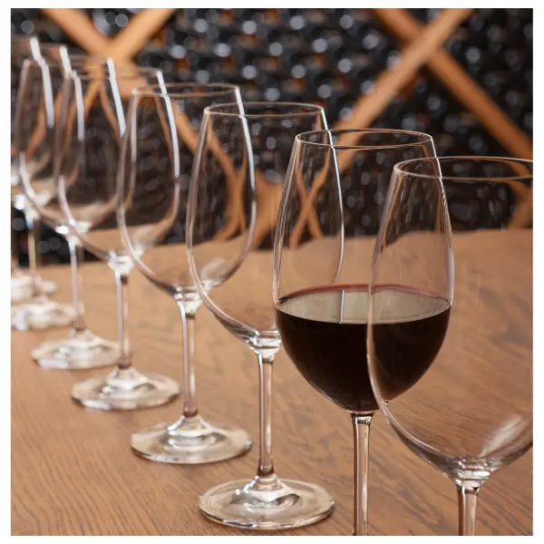 A number of wine glasses, one with red wine in it, lined up with wine bottles on shelves in the background
