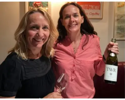 Two women at a tasting, one holding a glass and one showing a bottle of wine to the camera