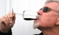 A man with grey hair and a beard wearing dark glasses and drinking some red wine